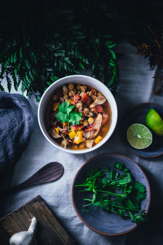 a flatlay top shot of a white bowl filled with vegan jackfruit chili alongside cilantro limes and framed by plants in dark moody food photography style