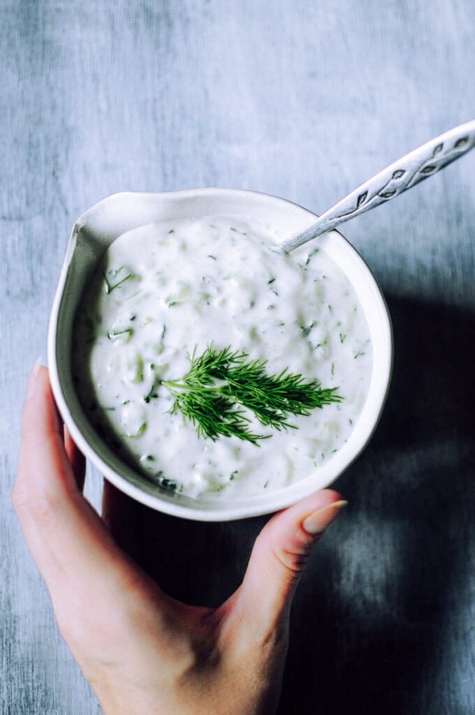 A hand holds a small bowl of tzatziki sauce topped with fresh green herbs.