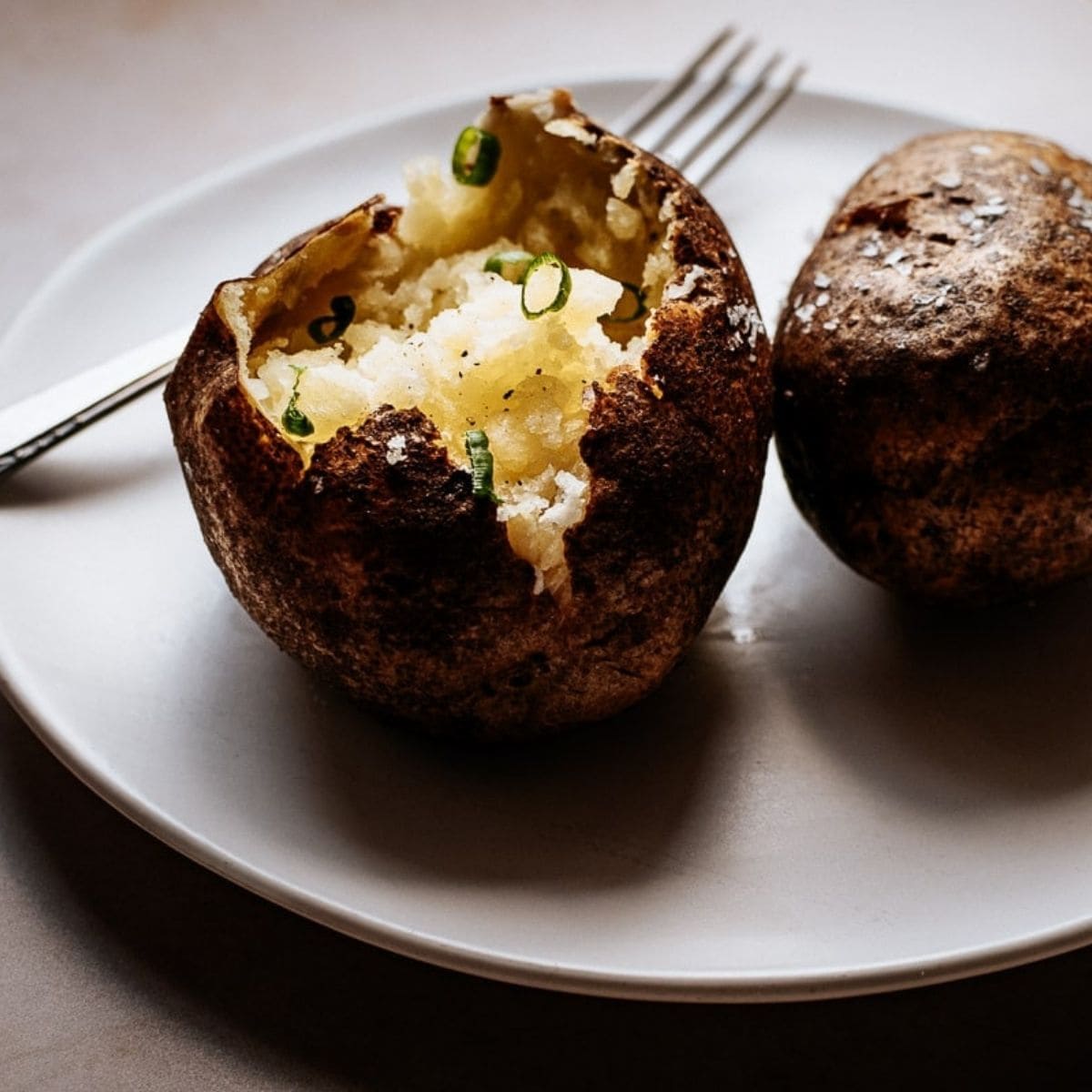 Two air fryer baked potatoes sit on a plate; one is cut open and topped with melting butter and fresh chives, with a fork resting beside them.