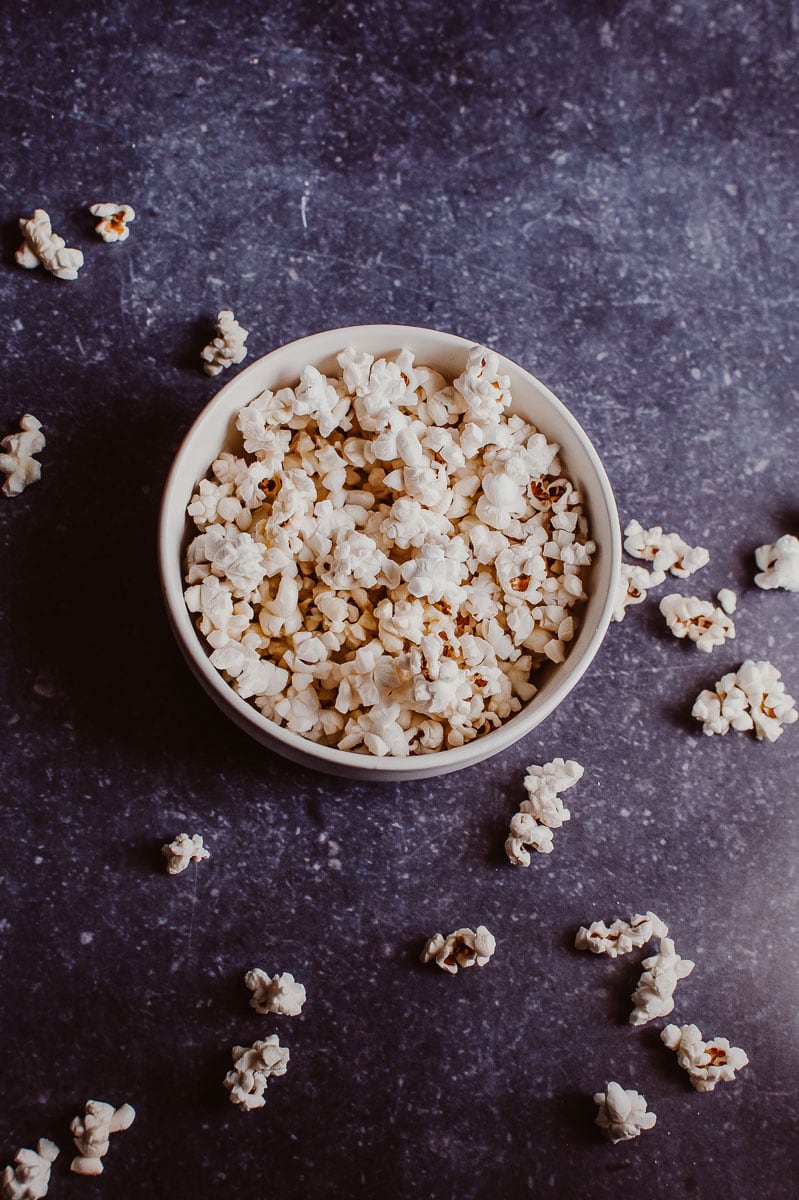 A white bowl filled with white popcorn overflowing onto the dark blue background.