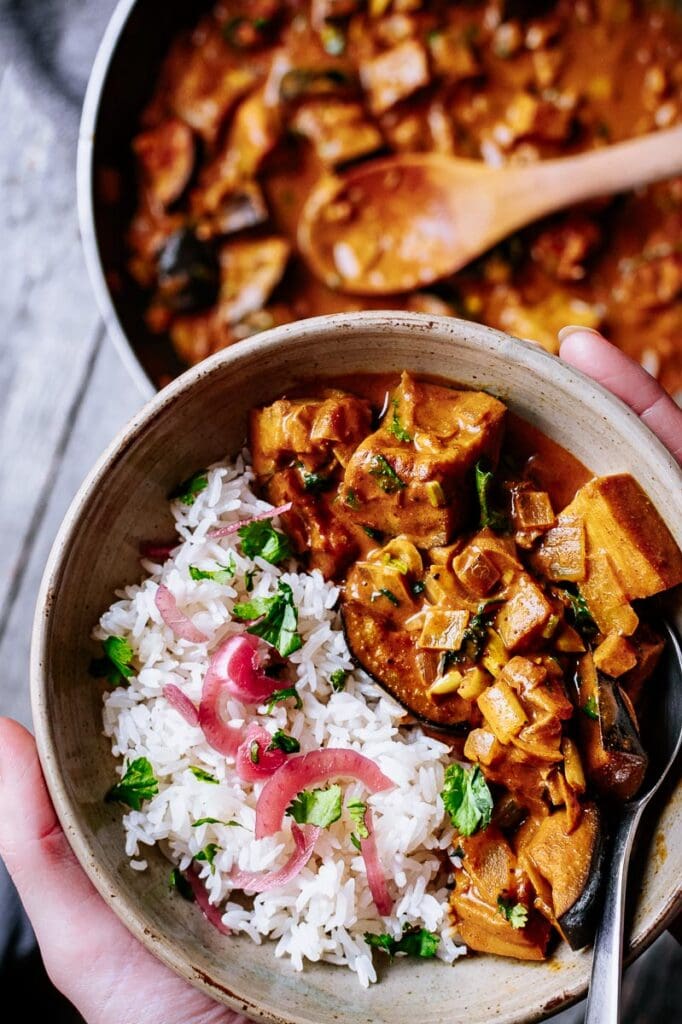 hands holding a bowl of orange red curry with rice and cilantro