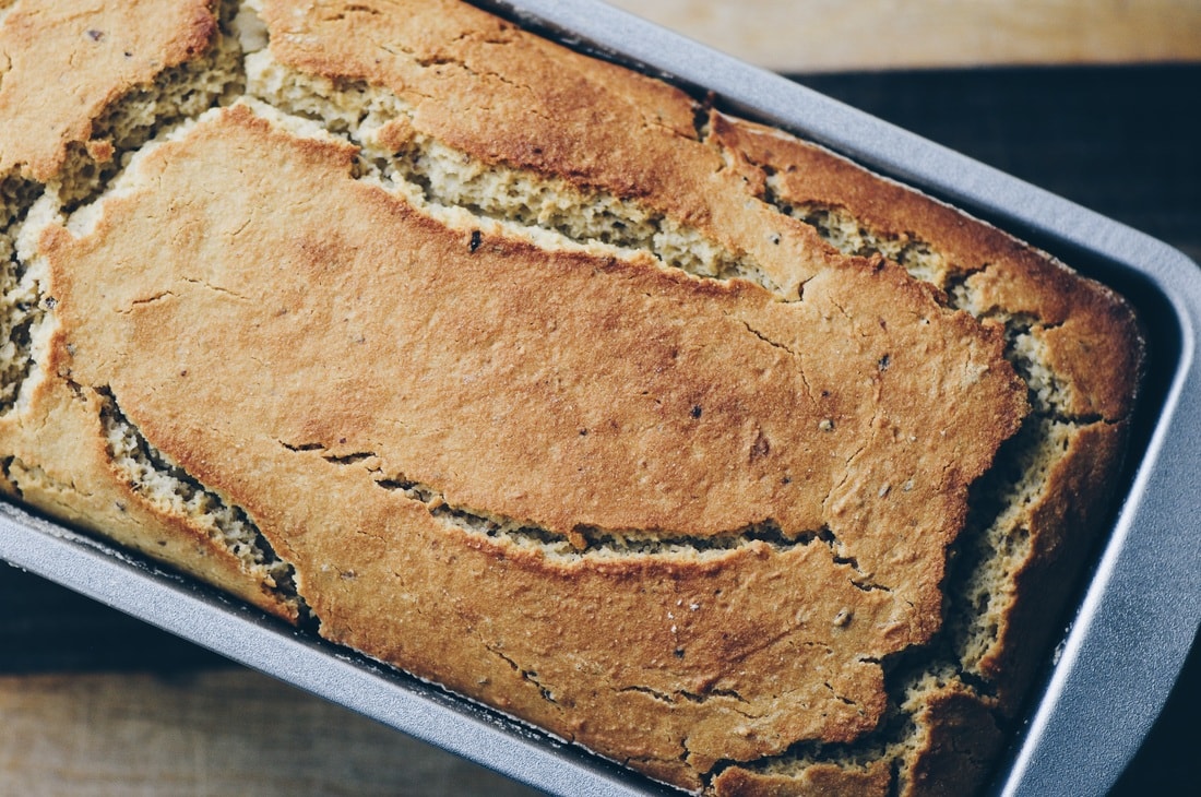 A loaf of golden-brown bread with a cracked top sits in a metal baking pan on a wooden surface.