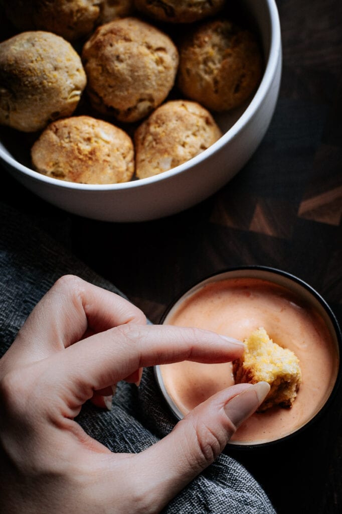 a hand holding a half bitten hush puppy about to be dipped into a red sauce