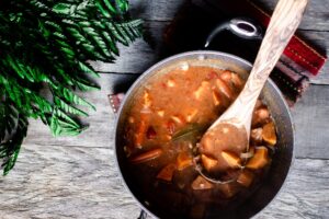 A pot of vegetarian stew with a wooden serving spoon, set on a rustic wooden table next to a fern frond.