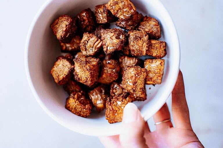 A hand holding a white bowl filled with browned, cubed pieces of air fryer tempeh.