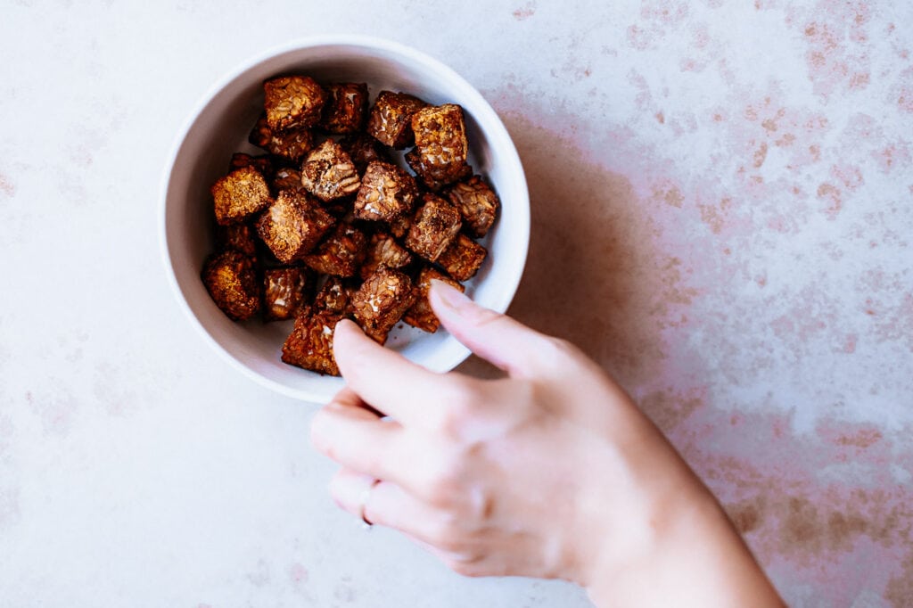 a hand reaching for a piece of tempeh in a white bowl