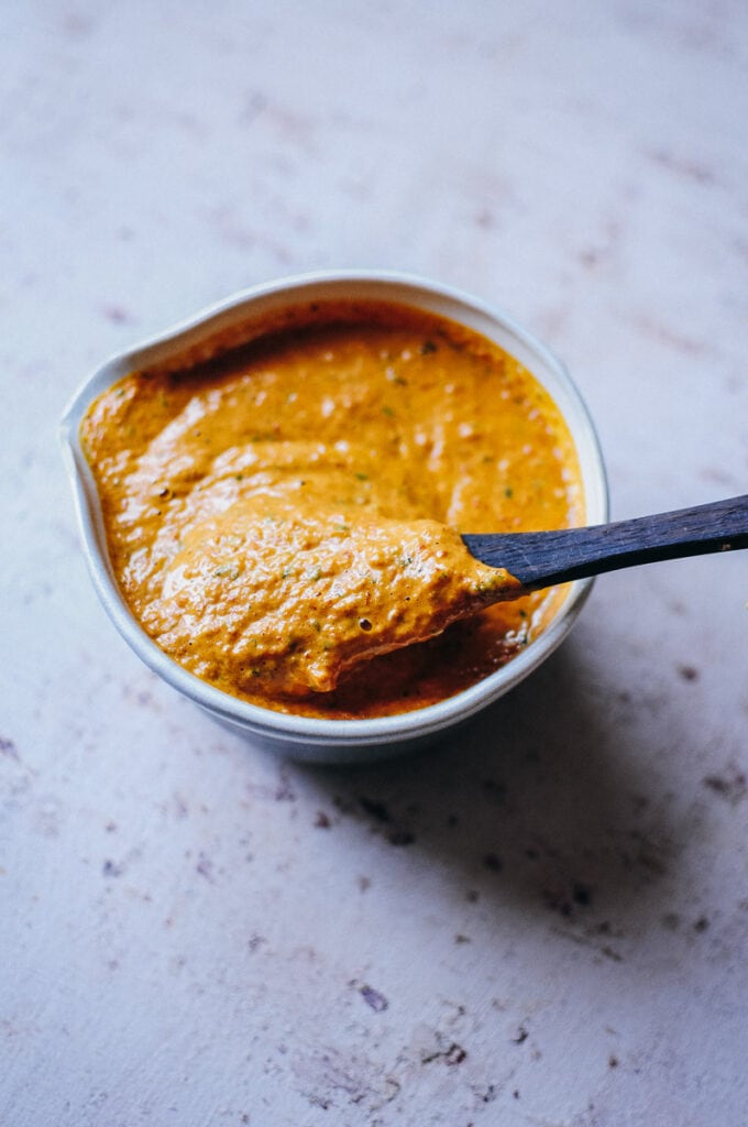 a wooden spoon scooping out almond butter romesco sauce from a ceramic bowl