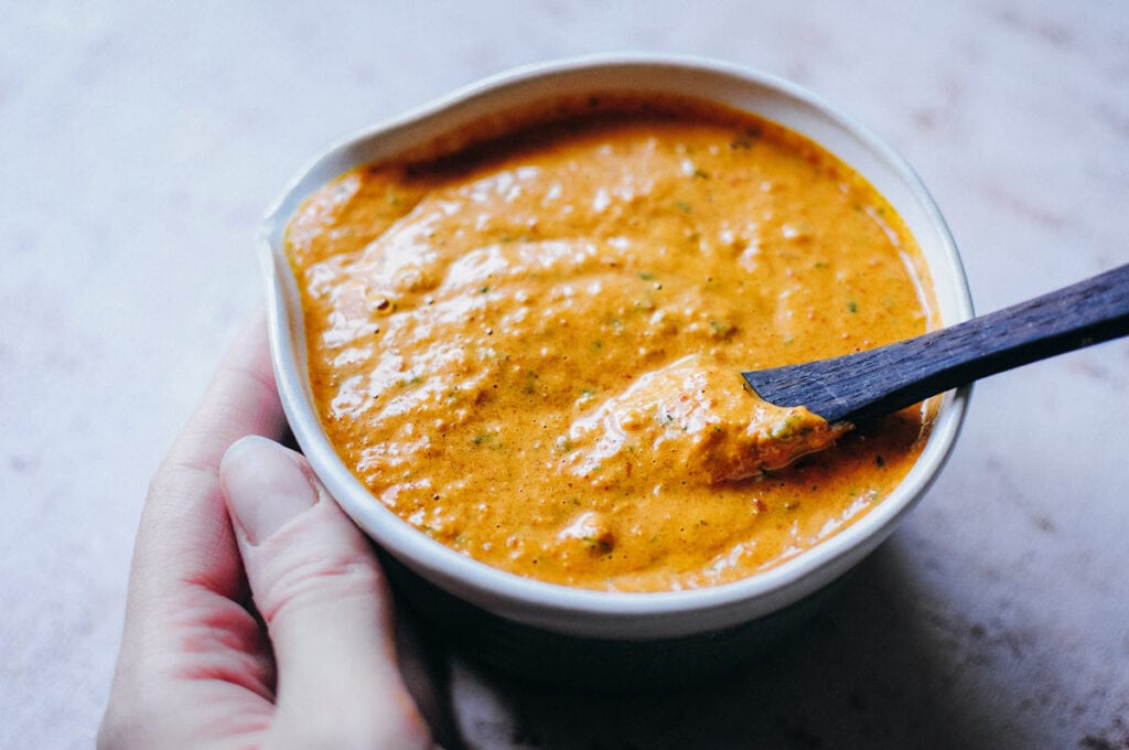 a hand holding a bowl of vibrant vegan romesco sauce