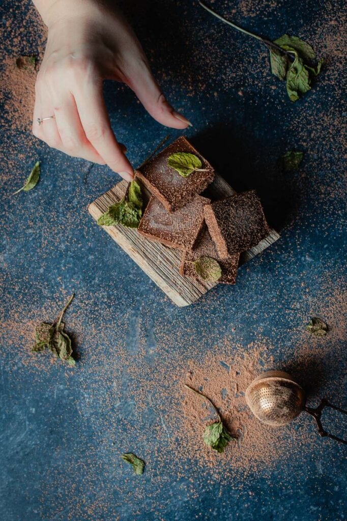 a hand reaching for a piece of mint fudge resting on a block of cut wood sprinkled with cocoa powder