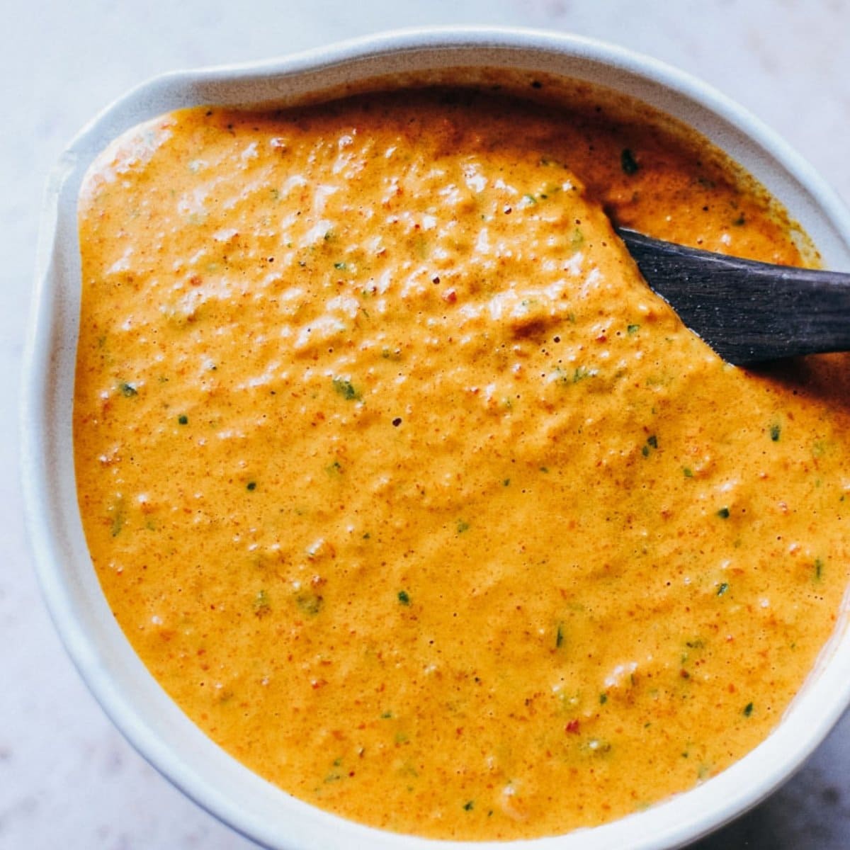 A close-up of a bowl filled with creamy, orange-colored romesco sauce with visible green and red specks. A black spoon rests inside the bowl, ready to scoop up this easy romesco sauce perfect for dipping or spreading.