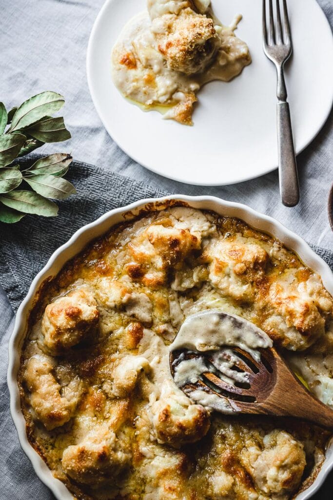 A wooden spoon resting in a large round white dish of gluten free cauliflower cheese.