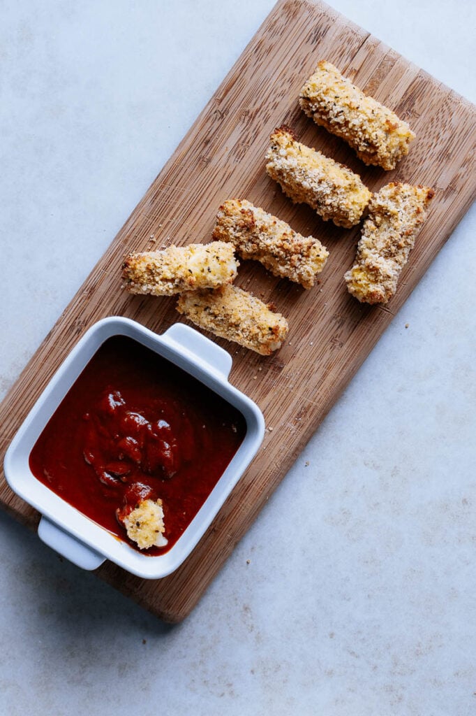a top view of a pile of gluten free air fryer mozzarella sticks on cutting board next to a white bowl of marinara dipping sauce