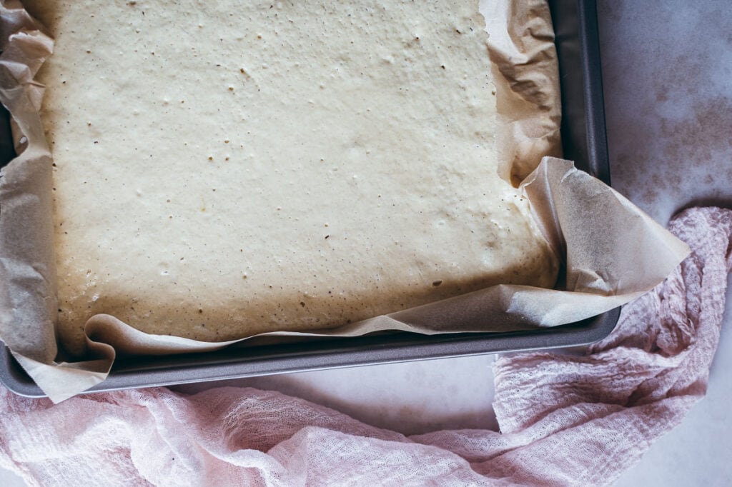 A parchment-lined baking pan filled with uncooked gluten free focaccia batter sits on a countertop next to a pink kitchen towel.