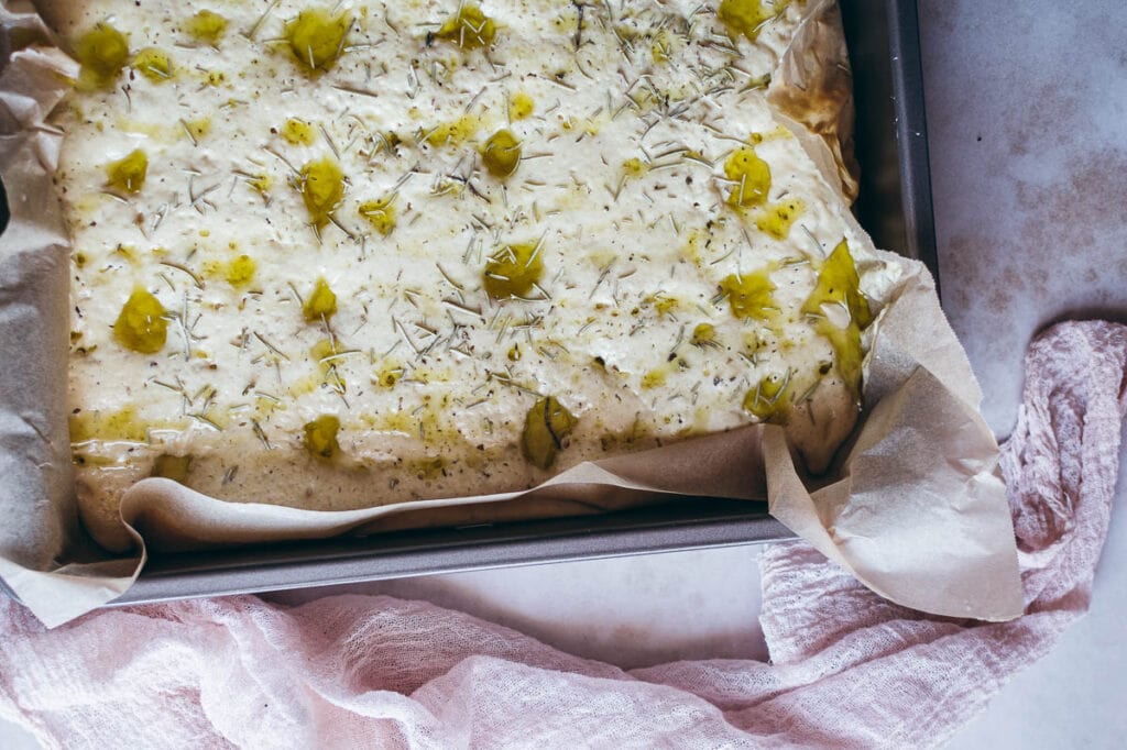 A close-up of unbaked gluten free focaccia dough in a parchment-lined baking pan, topped with herbs and olive oil, on a light surface with a pink cloth nearby.