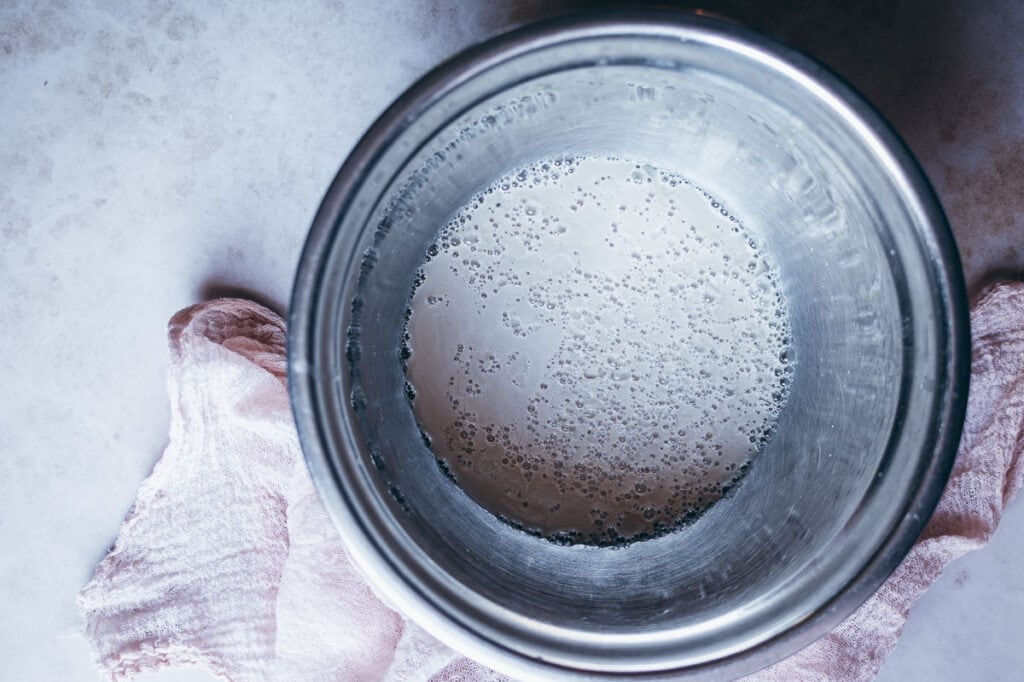 A metal bowl with a yeast mixture for gluten free focaccia bread sits on a pink cloth on a light surface, viewed from above.