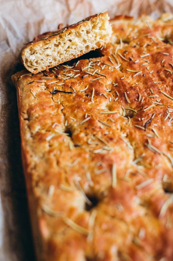 a slice of gluten free focaccia resting on top of the loaf of gf focaccia bread topped with rosemary and olive oil
