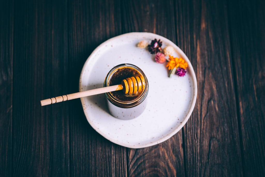 a white ceramic plate on a dark wooden table with an open jar of honey and dried wildflowers resting on it