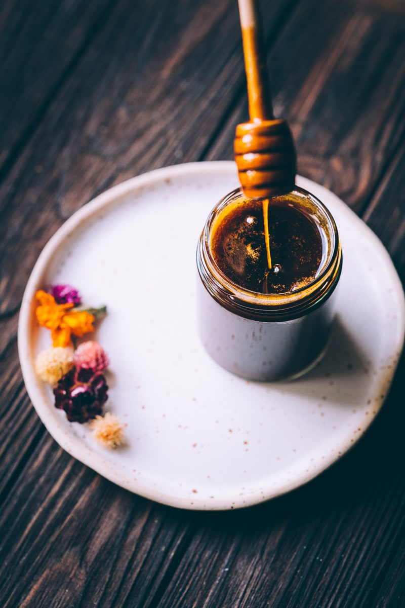 A wooden honey dipper drizzling honey into an open clear jar.