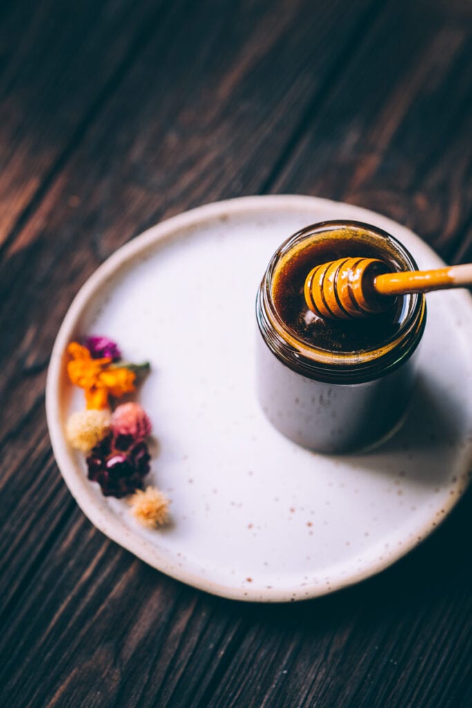 An open jar of dark honey rests on a speckled ceramic plate next to colorful dried flowers.
