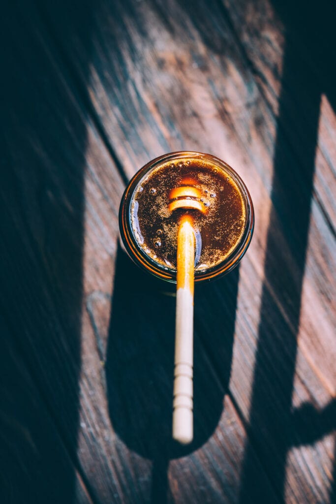 Sunlight hits a open jar filled with dark substance and topped with a wooden honey dipper stick.