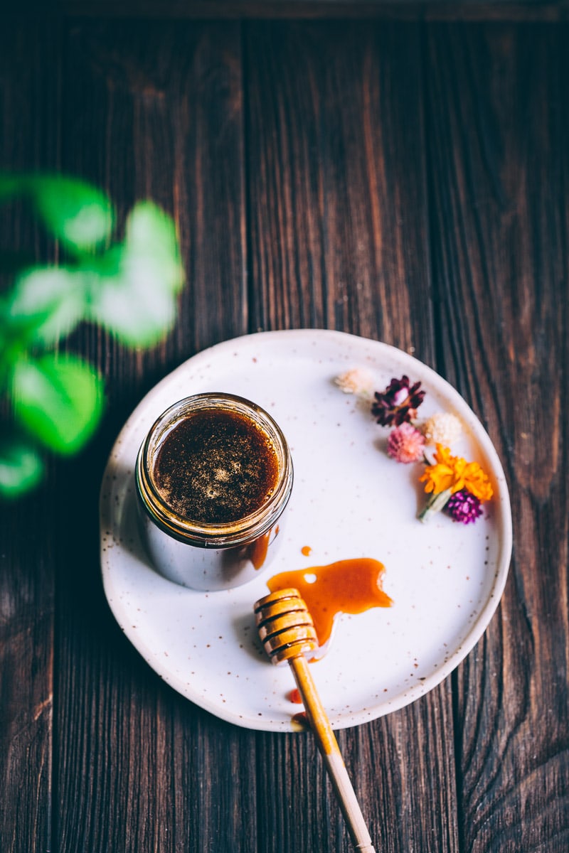 A jar of a dark liquid on a dark wooden table framed by green plants.