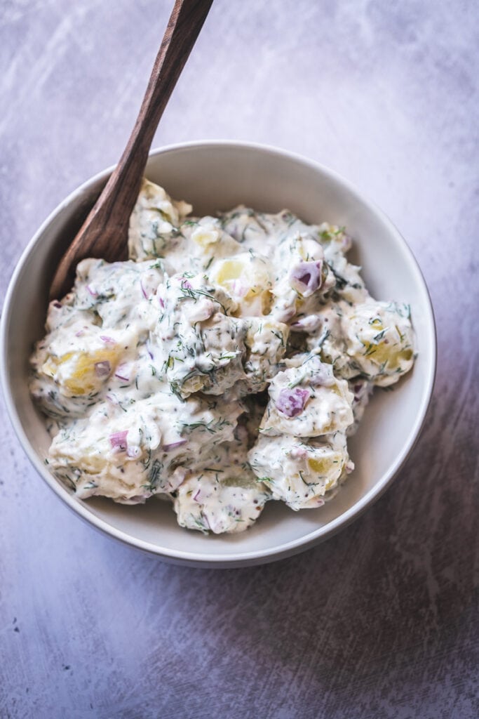 a wooden bowl sticks out of a white bowl of fresh dill potato salad