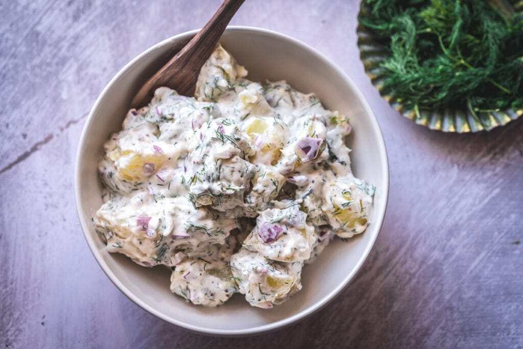 a white bowl filled with creamy potato salad alongside a dish of fresh dill