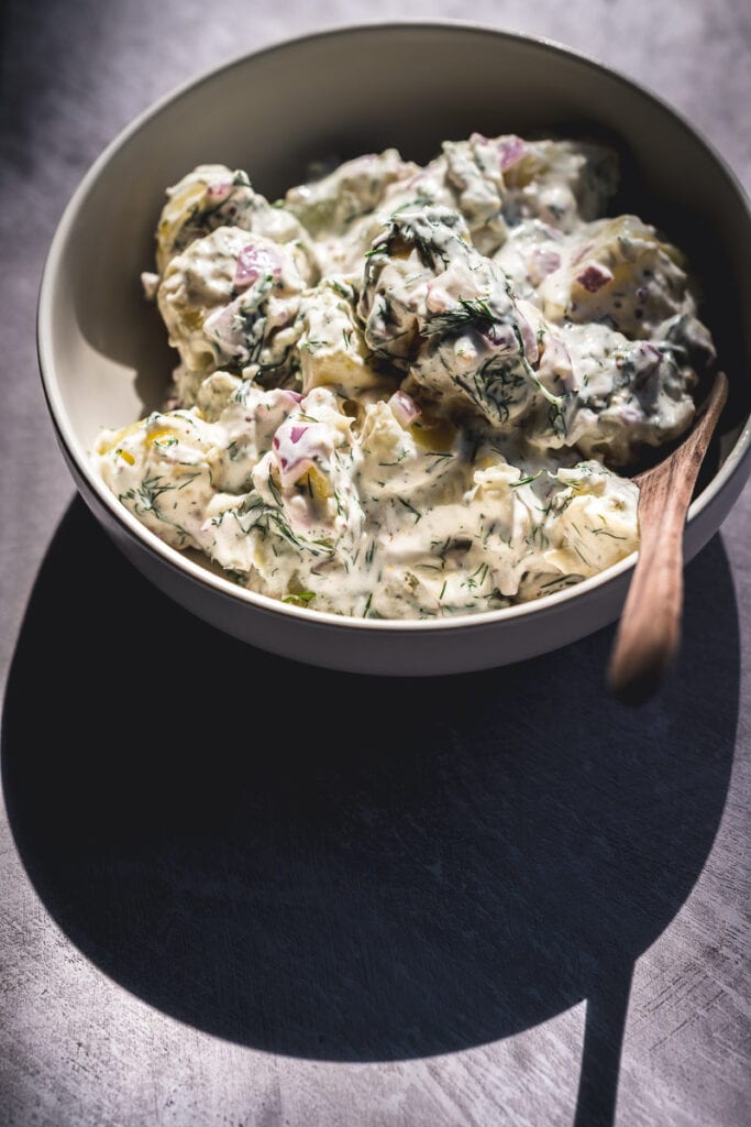 a bowl of creamy dill potato salad casts a long shadow on a grey backdrop