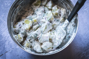 A metal bowl filled with creamy potato salad made from a classic dill potato salad recipe, mixed with red onion and fresh dill, with a black spoon inside.