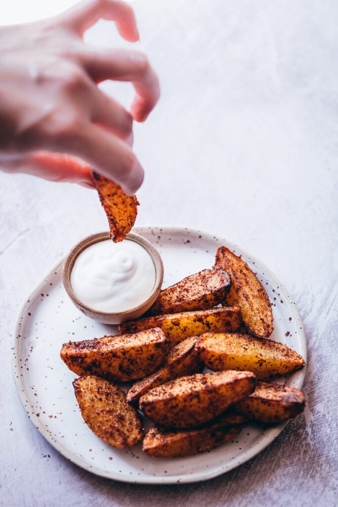 a hand holding an air fryer potato wedge about to dip in sour cream on a ceramic plate