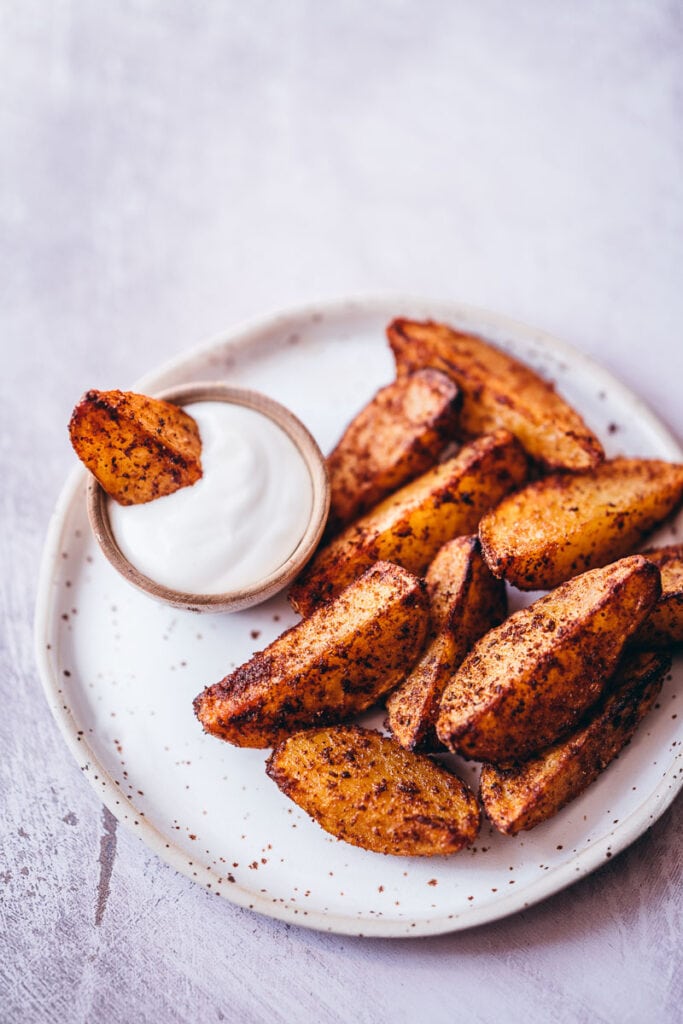 red seasoned smoky air fryer potato wedges sitting in a pile on a ceramic plate with one dipped in a small wooden bowl of white sour cream