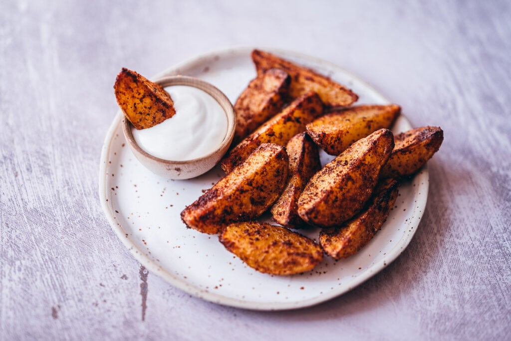 a ceramic plate topped with potato wedges with one dipped in a small wooden bowl of sour cream