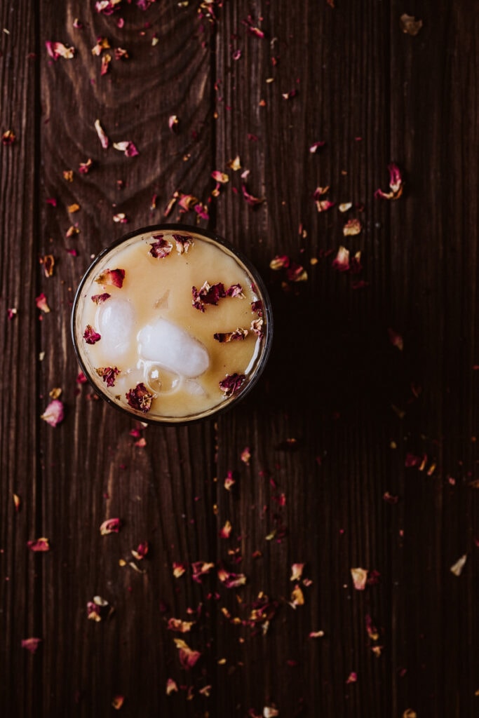 an iced rooibos tea latte on a dark wooden table topped with rose petals