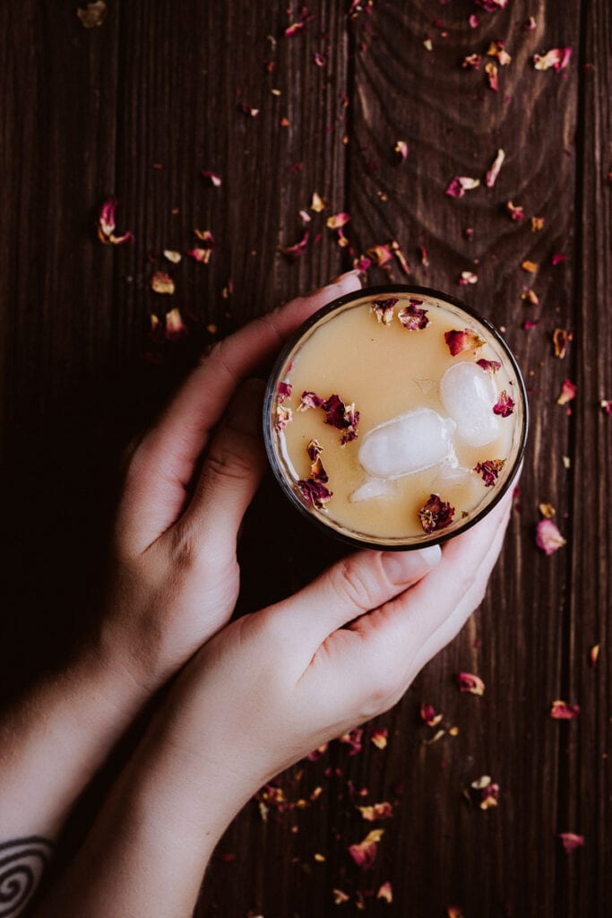 a hand holding a clear glass of iced rooibos scattered with rose petals