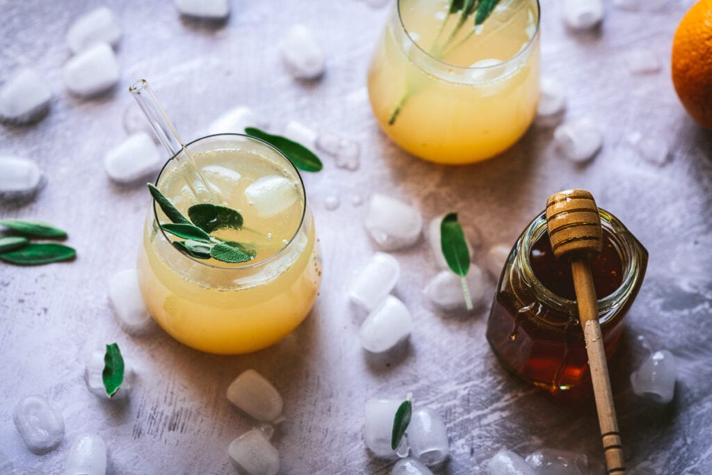 two clear glasses resting on ice cubes filled with mango orange shrub vinegar syrup recipe garnished with fresh sage and a glass straw