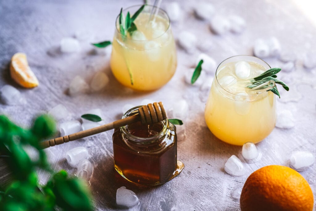 a wooden honey dipper dripping with orange honey alongside a fresh mango orange slices ice cubes and glasses of shrub vinegar drinks