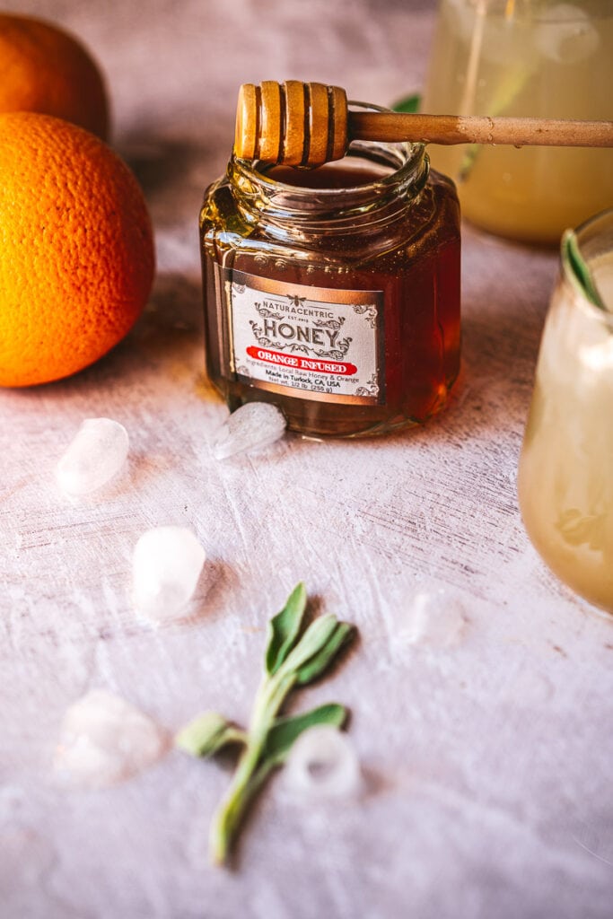 a glass jar of naturacentric orange infused honey with a wooden honey dipper resting on top dripping with orange honey