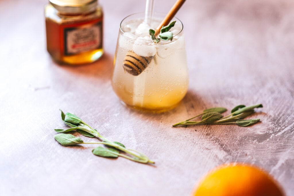a clear glass filled with orange shrub syrup and sparkling water with a wooden honey dipper stuck in