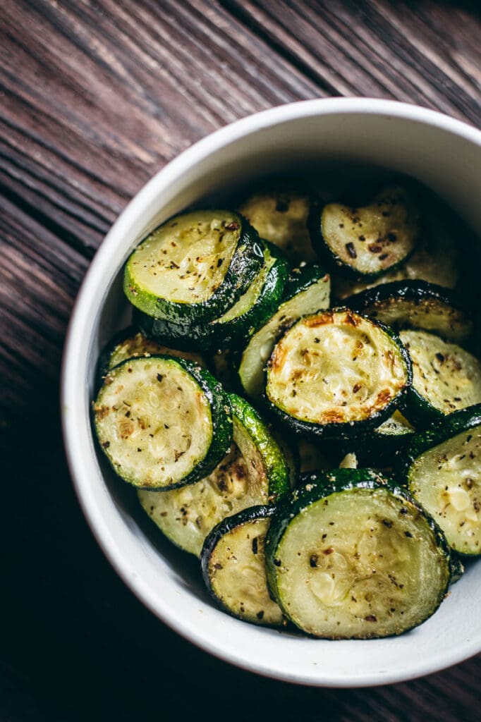 a close up shot of white bowl filled with zucchini slices cooked in the air fryer