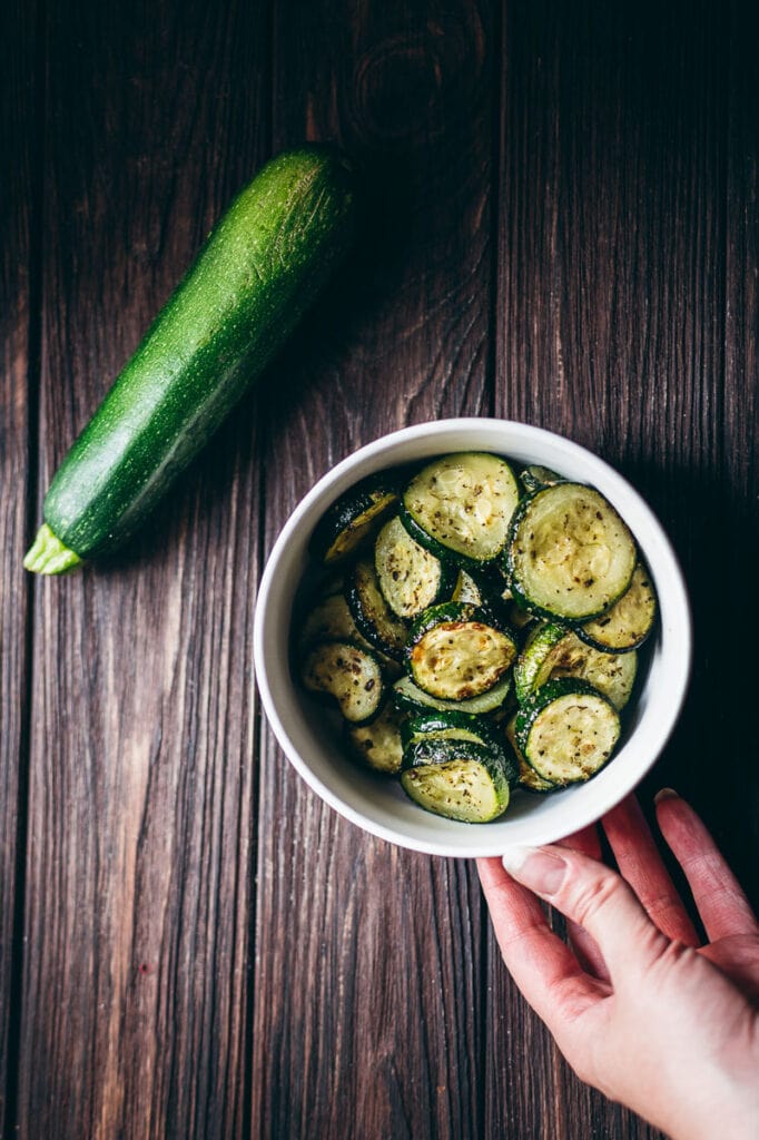 a white bowl of air fried zucchini resting on a wooden table next to a fresh green zucchini