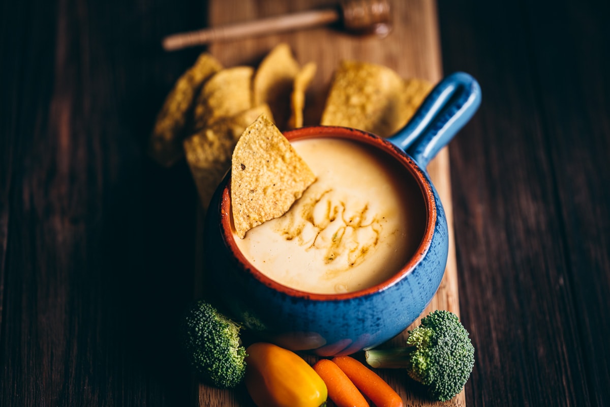 a blue ceramic crock filled with creamy spicy queso dip garnished with corn tortilla chips and fresh veggies