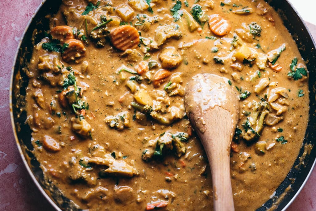 a skillet filled with orange veggie korma curry with a wooden spoon