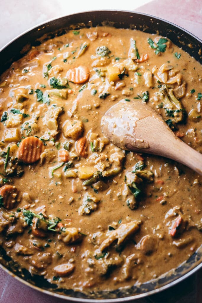 a wooden spoon stirs a giant pan of vegetables in a creamy golden sauce