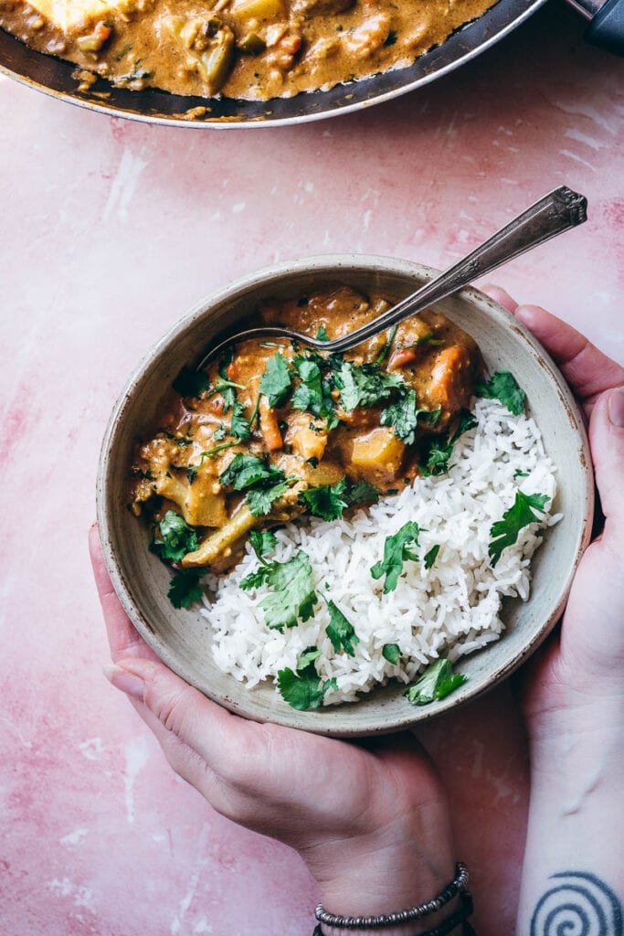 hands hold a gray ceramic bowl filled with veggie korma and white rice sprinkled with green cilantro