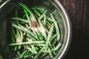 A metal bowl containing air fryer green beans—frozen green beans sprinkled with salt, pepper, and seasonings—rests on a dark wooden surface.