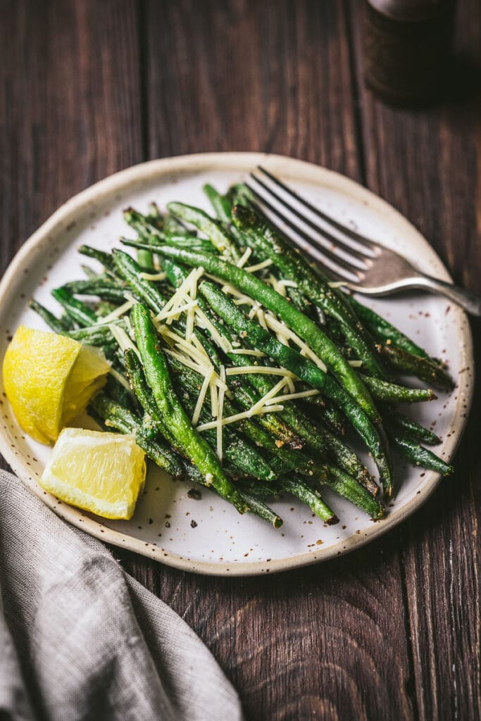 A plate filled with cooked french green beans lemon wedges and shredded parmesan cheese.