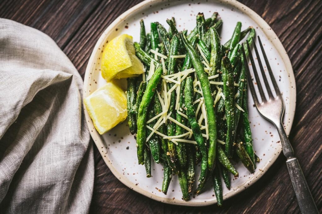 A white ceramic plate topped with crispy green beans made in the air fryer and topped with lemon and parmesan cheese.
