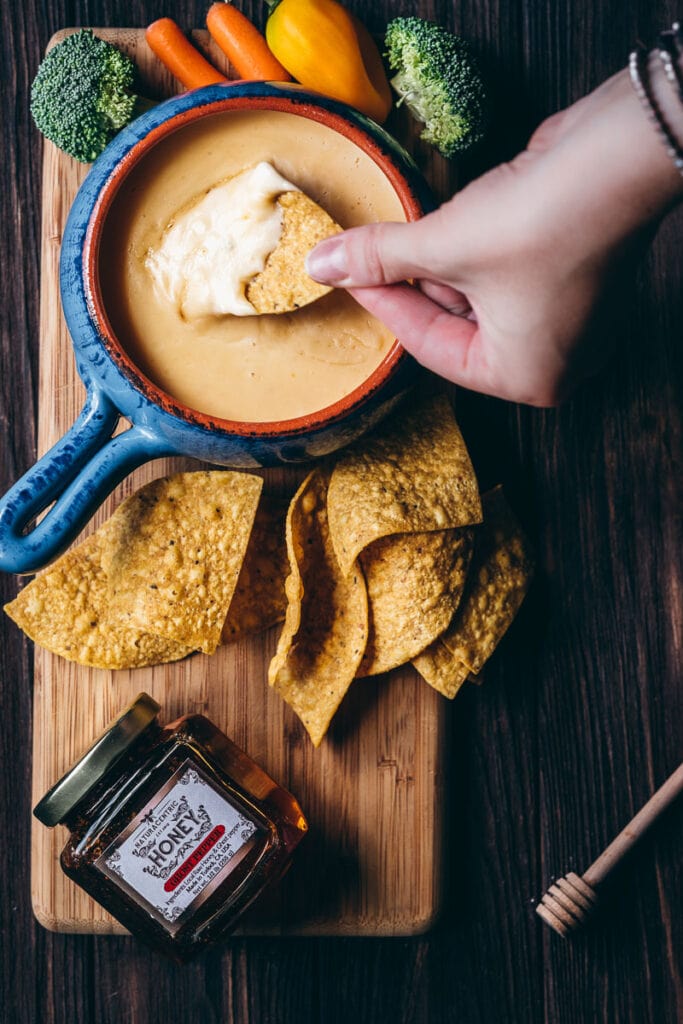 a hand dips a tortilla chip into a spicy gluten free queso dip made with ghost pepper honey