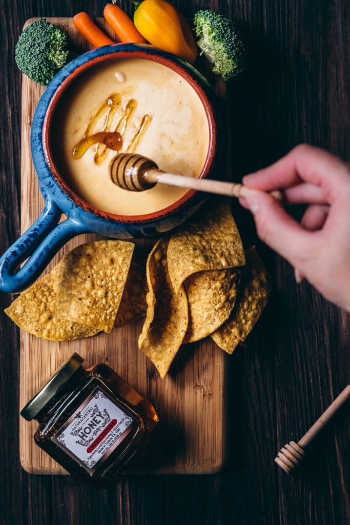 a hand drizzled ghost pepper infused honey a blue ceramic crock of gluten free spicy ghost pepper queso dip recipe