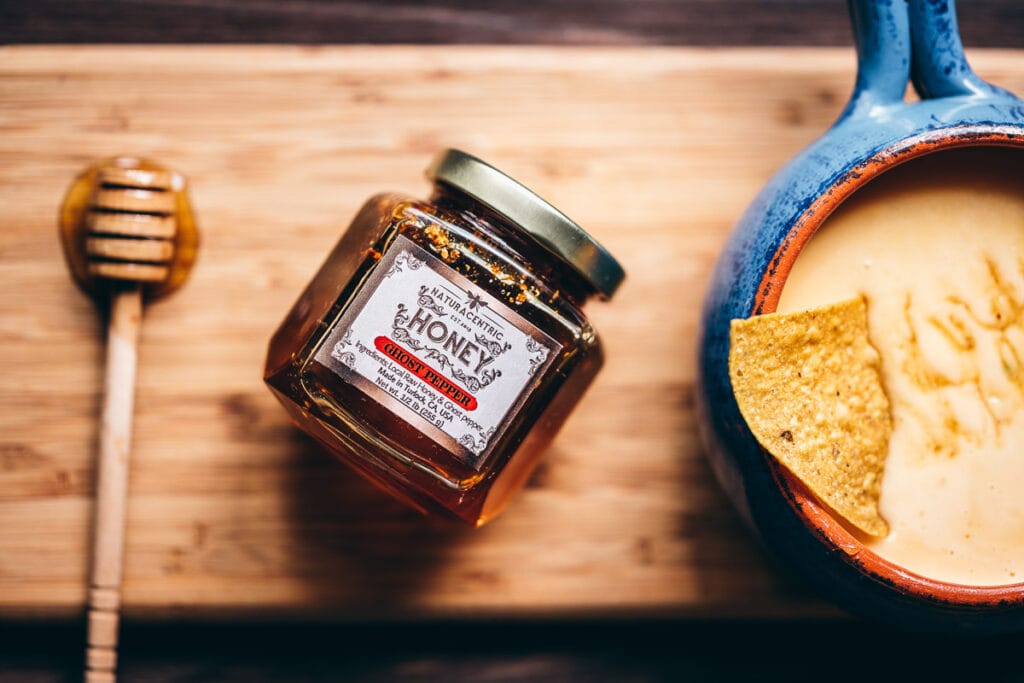 a honey spoon rests on a wooden cutting board next to a small jar of ghost pepper honey and a bowl of spicy queso dip